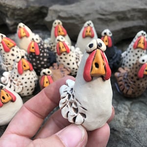 May include: A collection of hand-painted ceramic chickens, each with a unique personality. The chickens are white, black, and brown with red and orange accents. They are all standing on a rocky surface.