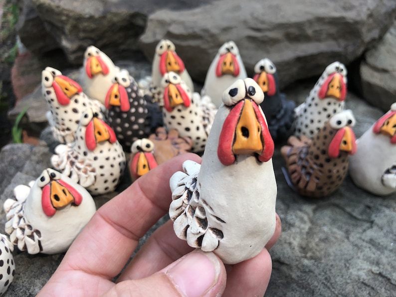 May include: A collection of hand-painted ceramic chickens, each with a unique personality. The chickens are white, black, and brown with red and orange accents. They are all standing on a rocky surface.