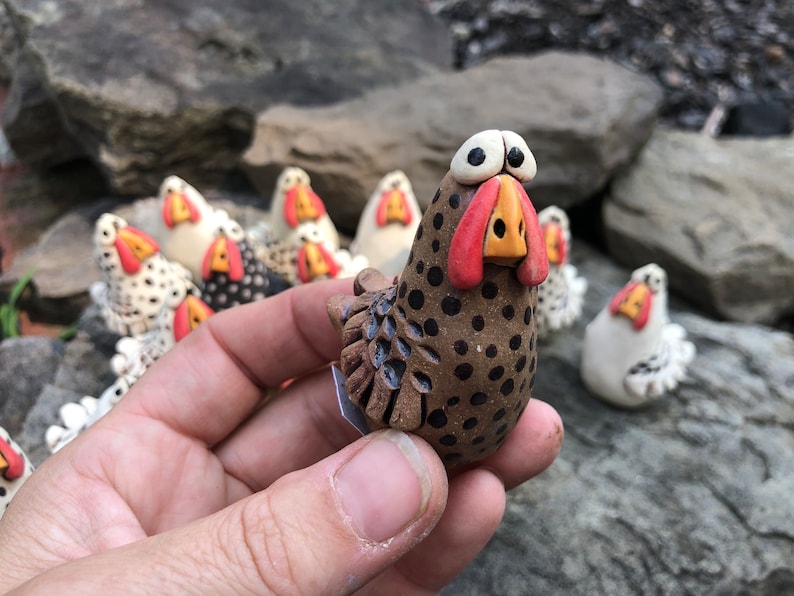 May include: A brown and black speckled ceramic chicken figurine with red and yellow accents. The chicken has large, round eyes and a wide, open beak. The figurine is about 3 inches tall and is standing on a small, flat base.