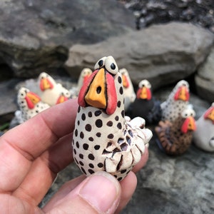 May include: A hand holds a small, white ceramic chicken figurine with black polka dots. The chicken has a red and yellow beak and large, black eyes. The figurine is surrounded by other similar figurines.