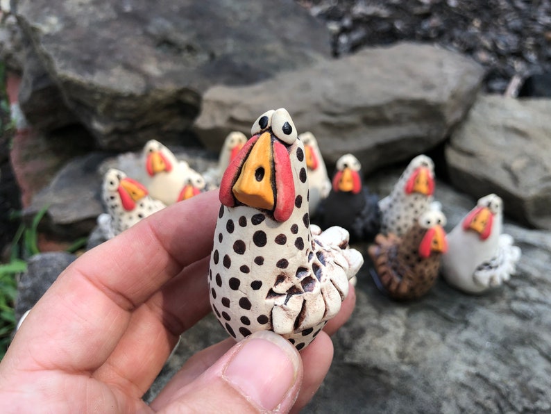 May include: A hand holds a small, white ceramic chicken figurine with black polka dots. The chicken has a red and yellow beak and large, black eyes. The figurine is surrounded by other similar figurines.