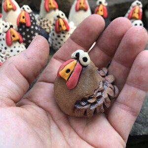 May include: A small, brown ceramic chicken figurine with large, white eyes and a red beak. The chicken has a detailed, textured body and is sitting on a hand.