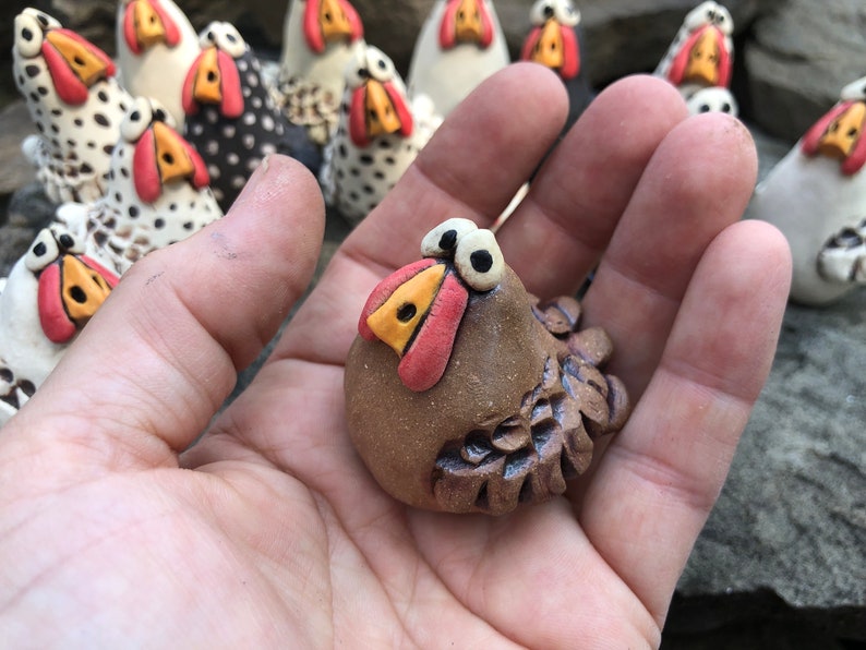 May include: A small, brown ceramic chicken figurine with large, white eyes and a red beak. The chicken has a detailed, textured body and is sitting on a hand.