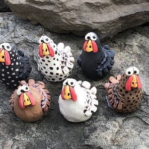 May include: Six ceramic chickens with different colored and patterned bodies. The chickens have large eyes, red beaks, and yellow feet. They are arranged in a semi-circle on a gray rock surface.