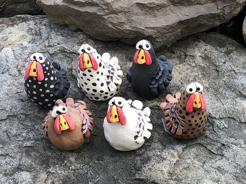 May include: Six ceramic chickens with different colored and patterned bodies. The chickens have large eyes, red beaks, and yellow feet. They are arranged in a semi-circle on a gray rock surface.