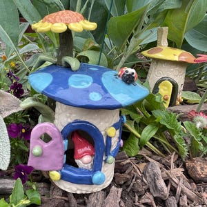 May include: Ceramic mushroom house with a blue spotted roof, a purple door, and a gnome inside. A ladybug sits on the roof. Another mushroom house with a yellow roof is in the background. The houses are surrounded by flowers and greenery.