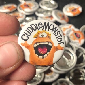 May include: A close-up of a round button with a white background and a cartoon orange monster with a wide open mouth and a pink tongue. The monster is wearing a red and yellow hat and has a yellow smile. The text "Cuddle Monster" is written in black around the monster.