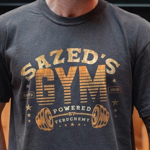 K&ouml;nnte beinhalten: Dunkelgraues T-Shirt mit einem verblassten goldenen Aufdruck, der "SAZED'S GYM" &uuml;ber einer Hantel und die Worte "POWERED BY FERUCHEMY" zeigt. Das Shirt hat einen klassischen Rundhalsausschnitt und kurze &Auml;rmel.