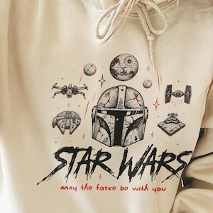 May include: A cream-colored hooded sweatshirt featuring a black and red graphic print of a Mandalorian helmet, various Star Wars spacecraft, and the text "STAR WARS" followed by the phrase "may the force be with you" in a handwritten red font.