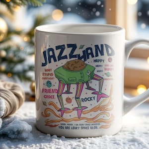 May include: White ceramic mug with the word "JAZZLAND" and a cartoon robot design. The mug features the phrases "Why Stupid Question?" and "You Are Leaky Space Blob." The mug is set against a snowy background with a Christmas tree.