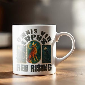 May include: White ceramic mug with the text "OMNIS LUPUS VIR" above "RED RISING" and a graphic of a red wolf. The mug has a curved handle and is set on a wooden surface. A coffee machine is in the background.