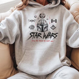 May include: A light gray hooded sweatshirt featuring a black and red graphic print of a Mandalorian helmet, various spacecraft, and the text Star Wars with the phrase May the force be with you. The hoodie includes a front kangaroo pocket and long sleeves.