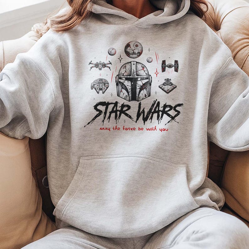 May include: A light gray hooded sweatshirt featuring a black and red graphic print of a Mandalorian helmet, various spacecraft, and the text Star Wars with the phrase May the force be with you. The hoodie includes a front kangaroo pocket and long sleeves.