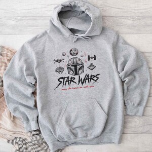 May include: A light gray hooded sweatshirt featuring a black and red graphic print of a Mandalorian helmet, various spacecraft, and the text "STAR WARS" with the phrase "may the force be with you" written in a stylized script below the main title.