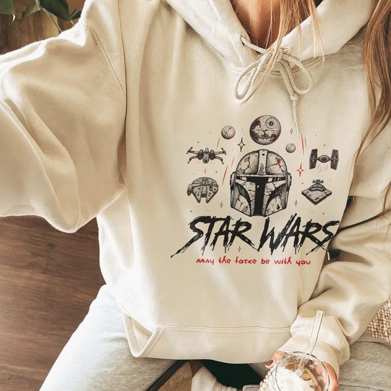 May include: A cream-colored hooded sweatshirt featuring a black and red graphic print of a Mandalorian helmet, various Star Wars spacecraft, and the text "STAR WARS" followed by the phrase "may the force be with you" in a handwritten red font.