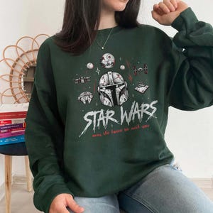 Op de afbeelding: Een donkergroen sweatshirt met lange mouwen en een grafische print van een Mandalorian-helm, de Death Star en diverse ruimteschepen. De tekst STAR WARS en may the force be with you staat in witte en rode letters op de borst.