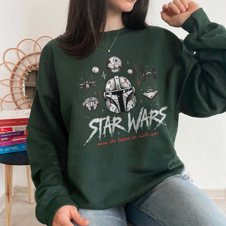 Op de afbeelding: Een donkergroen sweatshirt met lange mouwen en een grafische print van een Mandalorian-helm, de Death Star en diverse ruimteschepen. De tekst STAR WARS en may the force be with you staat in witte en rode letters op de borst.