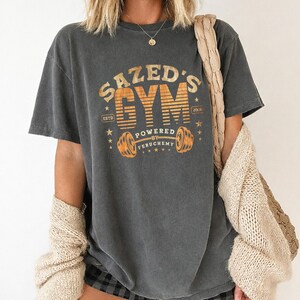 K&ouml;nnte beinhalten: Dunkelgraues T-Shirt mit dem Text "SAZED'S GYM" in Orange und Gelb. Das Shirt zeigt eine Hantel-Grafik und die Worte "POWERED BY FERUCHEMY". Das T-Shirt hat einen Vintage-Look.