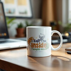 May include: White ceramic mug with the text "I Kept The Inside Thoughts Inside" in orange, green, and blue. The mug features a dragon and feather design. The mug is on a wooden desk with a laptop and notebook in the background.