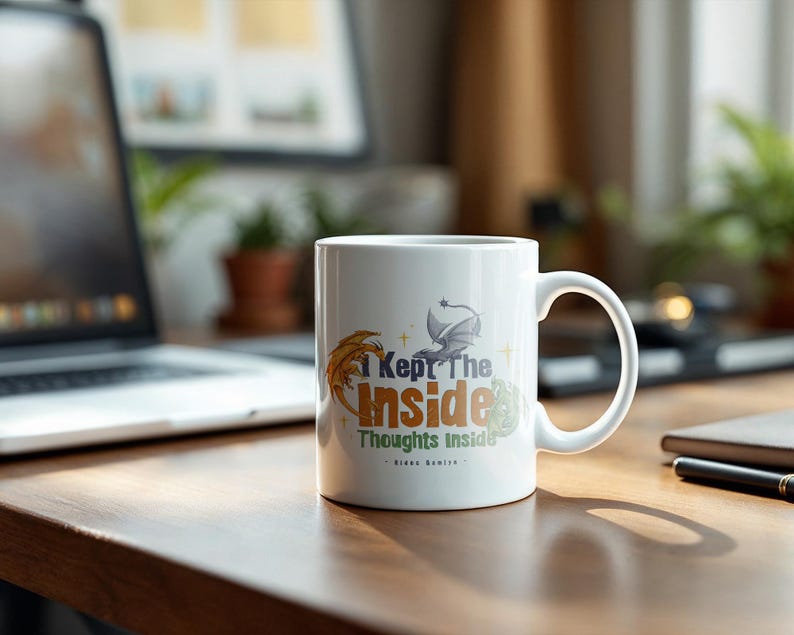 May include: White ceramic mug with the text "I Kept The Inside Thoughts Inside" in orange, green, and blue. The mug features a dragon and feather design. The mug is on a wooden desk with a laptop and notebook in the background.