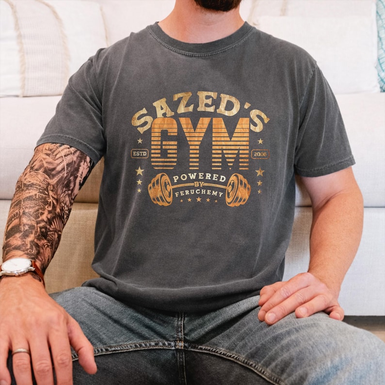 K&ouml;nnte beinhalten: Dunkelgraues T-Shirt mit einem verblassten goldenen Aufdruck, der "SAZED'S GYM" mit einer Hantel-Illustration zeigt. Das Shirt enth&auml;lt auch den Text "ESTD 2008" und "POWERED BY FERUCHEMY". Das T-Shirt wird von einer Person getragen.