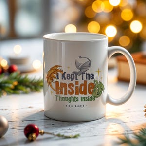 May include: White ceramic mug with a curved handle. The mug features a design with three dragons in orange, gray, and green, along with the text "I Kept The Inside Thoughts Inside". The mug is on a white wooden surface with Christmas decorations.