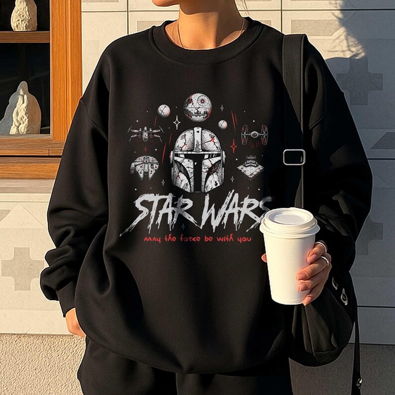 Op de afbeelding: Een zwart sweatshirt met ronde hals en een grafische print van een Mandalorian-helm, de Death Star en diverse ruimteschepen. De tekst STAR WARS en May the force be with you staat in witte en rode letters op de borst van het kledingstuk.