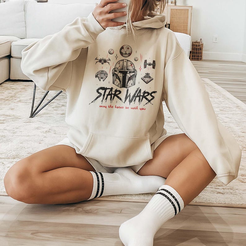 May include: A cream-colored hooded sweatshirt featuring a graphic print of a Mandalorian helmet, various Star Wars spacecraft, and the text "STAR WARS" in a distressed black font, with the phrase "may the force be with you" in red text below.