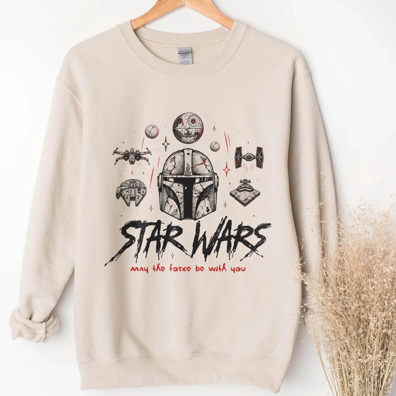 Op de afbeelding: Een beige sweater met ronde hals en een zwarte grafische print van een Mandalorian-helm, de Death Star, een X-wing, een TIE-fighter en de Millennium Falcon. De tekst luidt STAR WARS en May the force be with you in zwarte en rode inkt.