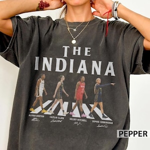 May include: A charcoal gray short-sleeve t-shirt featuring the text THE INDIANA in large white letters above four basketball players in uniform. Below each player are the names Aliyah Boston, Caitlin Clark, Kelsey Mitchell, and a signature.