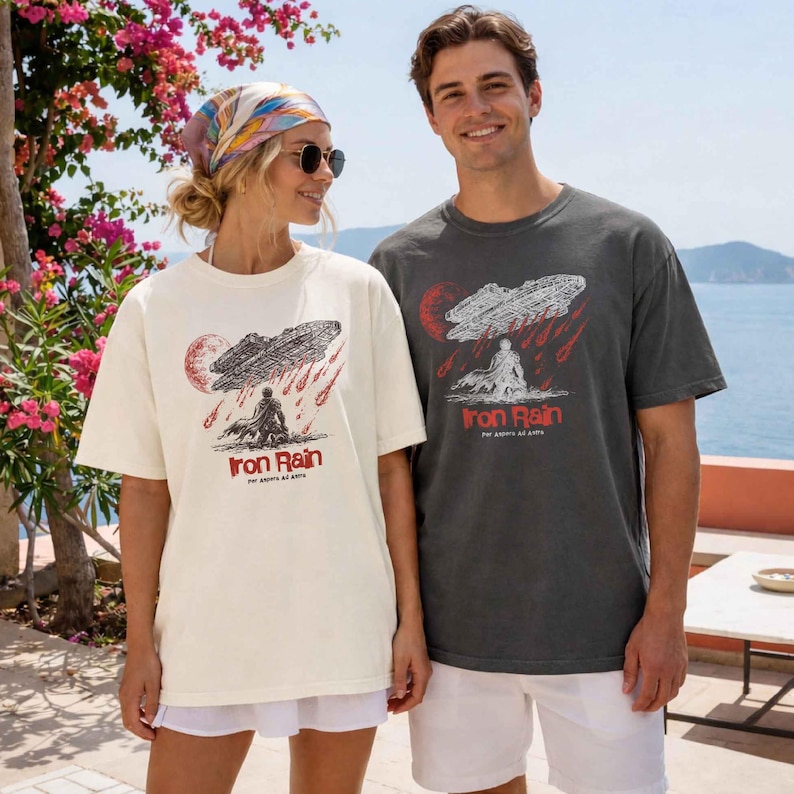 Op de afbeelding: Twee mensen dragen bijpassende t-shirts met een grafisch ontwerp. De shirts hebben een rood-zwarte illustratie van een figuur onder een wolk met de tekst "Iron Rain". De shirts zijn cr&egrave;me en antracietgrijs.