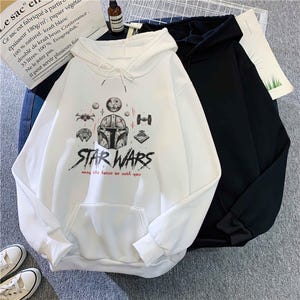 May include: A white hooded sweatshirt featuring a graphic print of a Mandalorian helmet, various spacecraft, and the text "STAR WARS" with "may the force be with you" in red. A black hooded sweatshirt is positioned partially behind the white one.
