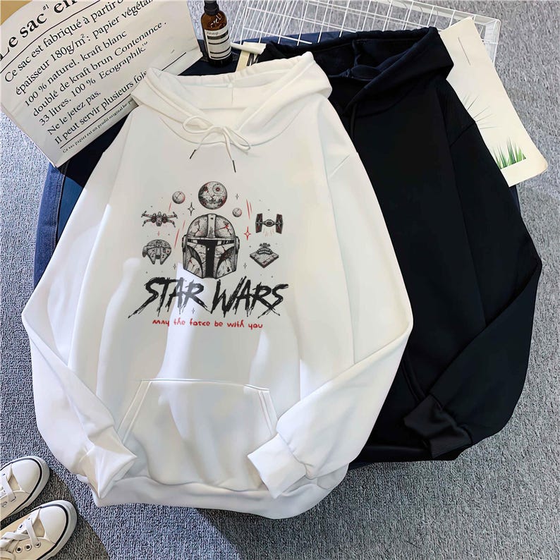 May include: A white hooded sweatshirt featuring a graphic print of a Mandalorian helmet, various spacecraft, and the text "STAR WARS" with "may the force be with you" in red. A black hooded sweatshirt is positioned partially behind the white one.
