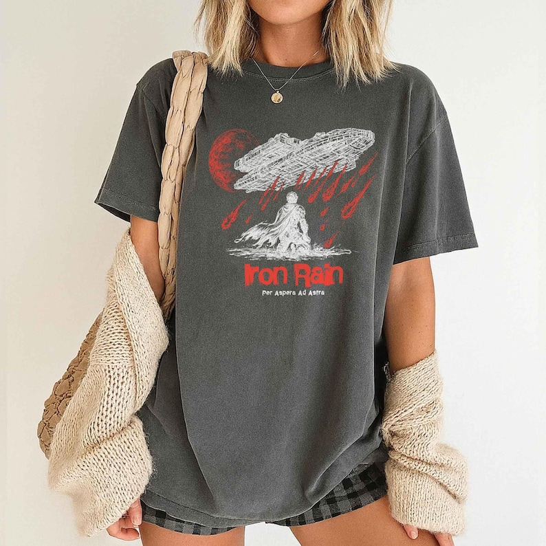Op de afbeelding: Donkergrijs t-shirt met een grafisch ontwerp. Het ontwerp bevat de tekst "Iron Rain" in het rood, een ruimteschip en een figuur. De zin "Per Aspera Ad Astra" staat eronder.