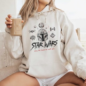May include: A cream-colored hooded sweatshirt featuring a black and red graphic print of a Mandalorian helmet, Star Wars logo, and various spacecraft. The text reads Star Wars and May the force be with you in a stylized black and red font.