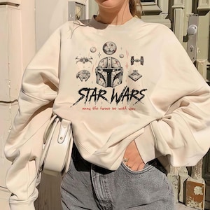 Op de afbeelding: Een cr&egrave;mekleurig sweatshirt met ronde hals voorzien van een grafische print van een Mandalorian-helm, diverse Star Wars-ruimteschepen en de tekst "STAR WARS" in het zwart met de zin "may the force be with you" in rode letters.