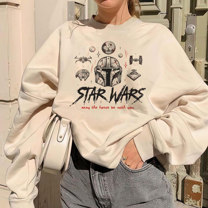 Op de afbeelding: Een cr&egrave;mekleurig sweatshirt met ronde hals voorzien van een grafische print van een Mandalorian-helm, diverse Star Wars-ruimteschepen en de tekst "STAR WARS" in het zwart met de zin "may the force be with you" in rode letters.