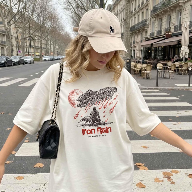 Op de afbeelding: Cr&egrave;me-wit T-shirt met een grafisch ontwerp met een ruimteschip, een figuur en de woorden "Iron Rain". Het shirt wordt gecombineerd met een beige baseballcap met een zwart logo en een zwarte schoudertas.