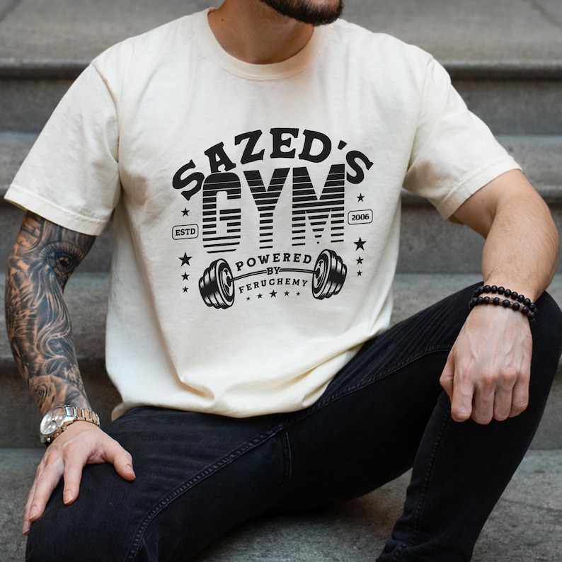K&ouml;nnte beinhalten: Cremefarbenes T-Shirt mit schwarzem Aufdruck &bdquo;SAZED'S GYM&ldquo; &uuml;ber einer Hantel-Illustration. Der Text &bdquo;ESTD 2006&ldquo; ist ebenfalls enthalten. Das T-Shirt wird von einer Person getragen, die auf Stufen sitzt.