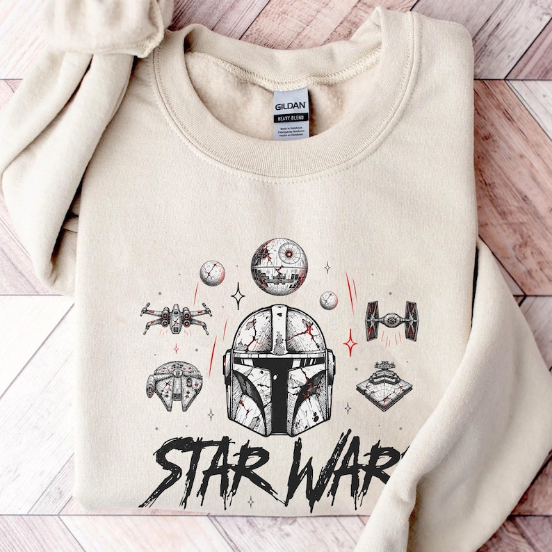 Op de afbeelding: Een zandkleurige sweater met ronde hals en een grafische print van een helm in Mandalorian-stijl, omringd door diverse Star Wars-ruimteschepen en de Death Star. De tekst STAR WARS staat in een verweerd zwart lettertype onderaan de print.