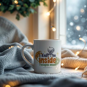 May include: White ceramic mug with the text "I Kept The Inside Thoughts Inside" and a dragon graphic. The mug has a curved handle and is set against a cozy, winter-themed background with soft lighting and a blanket.