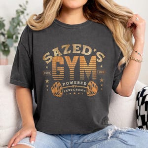 K&ouml;nnte beinhalten: Dunkelgraues T-Shirt mit einem Used-Design. Das Shirt zeigt den Text "SAZED'S GYM" in einem Vintage-Stil, zusammen mit einer Hantel-Grafik und den Worten "POWERED BY FERUCHEMY". Das Shirt enth&auml;lt auch den Text "ESTD 2006".