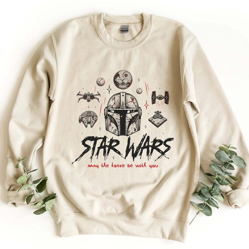 Op de afbeelding: Een beige sweater met ronde hals en een zwart-rode grafische print van een Mandalorian-helm, de Death Star, ruimteschepen en de tekst "STAR WARS" gevolgd door de zin "may the force be with you" in een gestileerd handgeschreven lettertype.