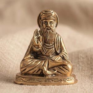May include: A small brass figurine of a bearded figure wearing a turban and robes, seated in a cross-legged position with one hand raised in a gesture of blessing. The base features an engraved pattern, and a small bird rests near the feet.