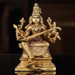 May include: A brass figurine of a deity seated on a rectangular base, holding a stringed musical instrument across the lap. The figure features intricate carvings, a tall crown, and multiple arms, with a reflective surface showing a clear reflection.