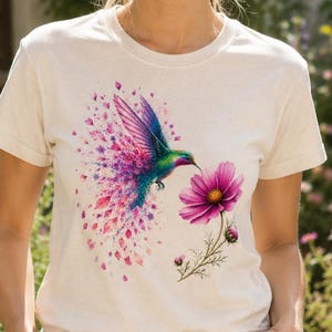 Peut inclure: Un t-shirt à manches courtes de couleur crème arborant une impression graphique d'un colibri vibrant aux plumes bleues et vertes irisées, entouré d'une traînée de pétales de fleurs roses et violettes, s'approchant d'une fleur cosmos rose vif.