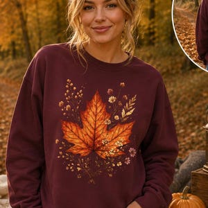 Könnte beinhalten: Ein kastanienbraunes Sweatshirt mit Rundhalsausschnitt, das auf der Vorderseite eine große, detaillierte Grafik eines orangefarbenen Ahornblatts umgeben von kleinen Wildblumen zeigt, sowie ein kleineres passendes Blumenmotiv auf dem oberen Rücken.