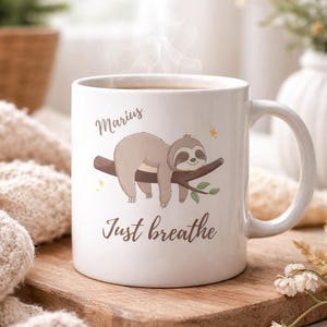 Könnte beinhalten: Weiße Keramiktasse mit einem Cartoon-Faultier auf einem Ast, dem Namen "Marius" und dem Schriftzug "Just breathe". Die Tasse steht auf einem Holzbrett. Dampf steigt auf, was auf ein heißes Getränk hindeutet.