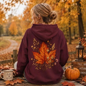 Könnte beinhalten: Ein kastanienbrauner Kapuzenpullover mit einer großen, detaillierten Grafik eines leuchtend orangefarbenen Ahornblatts, das von kleinen Wildblumen umgeben ist, auf der Rückseite. Der Hoodie hat lange Ärmel und eine Kapuze mit botanischem Motiv.
