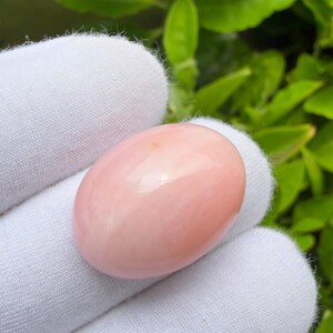 May include: An oval-shaped, smooth, light pink gemstone with a polished, glossy finish. The stone features a solid, opaque appearance with subtle variations in its soft pink hue, showcasing a rounded cabochon cut that reflects light evenly.